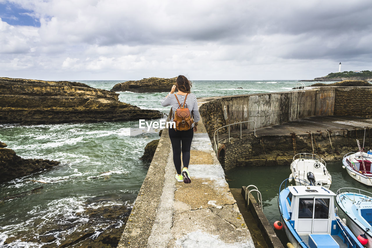 Rear view of young woman standing with backpack walking at beach against cloudy sky