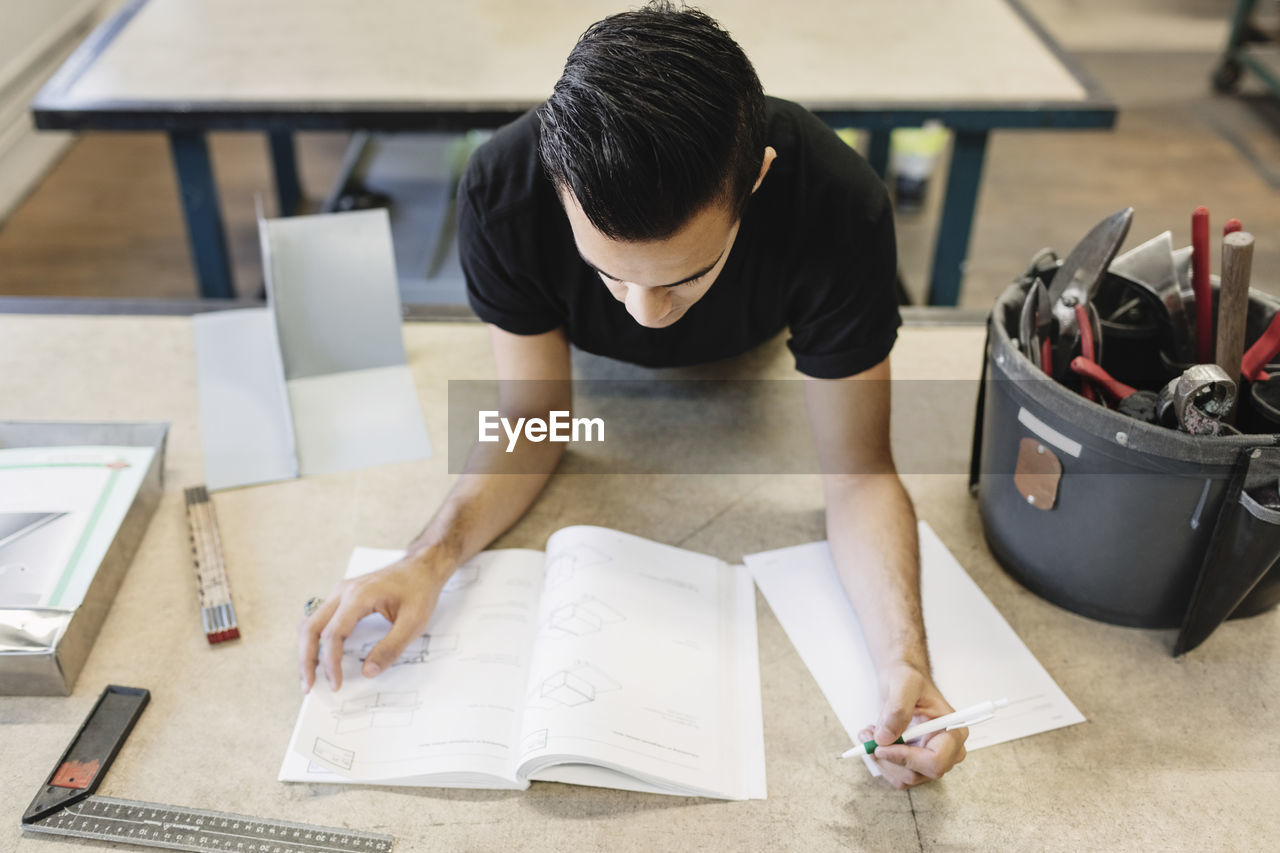 High angle view of male high school student writing in book at workshop