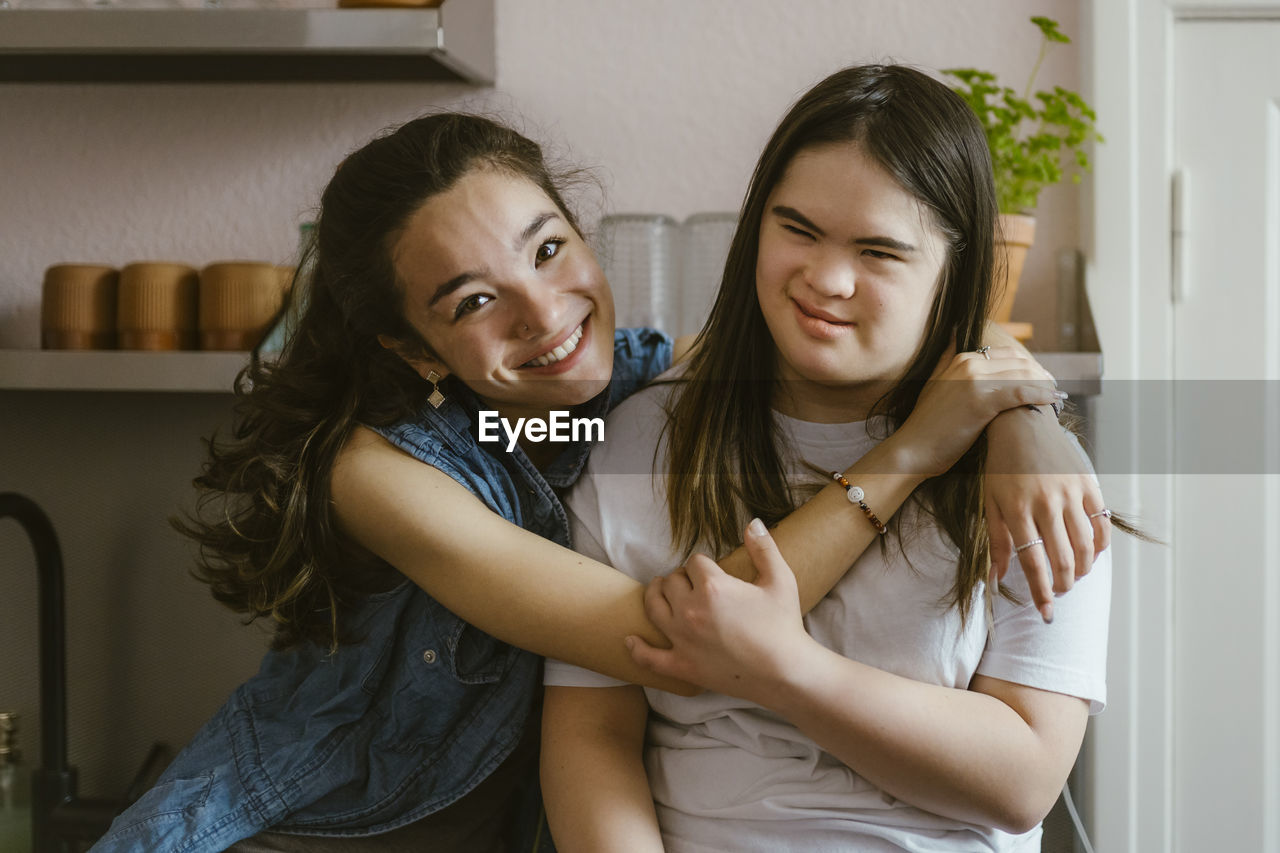 Portrait of smiling young woman hugging sister with down syndrome in kitchen at home