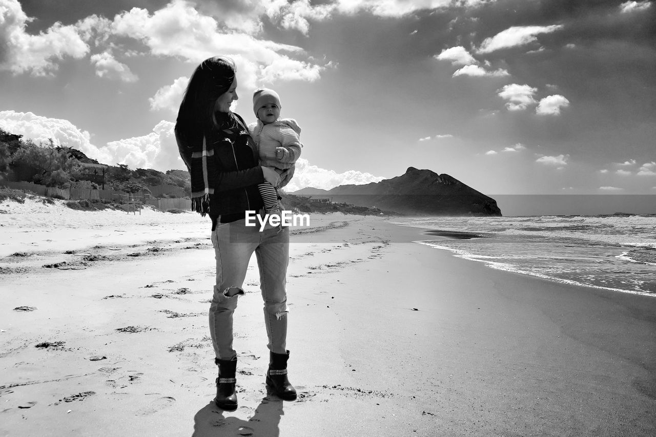 Mother carrying daughter at beach against sky