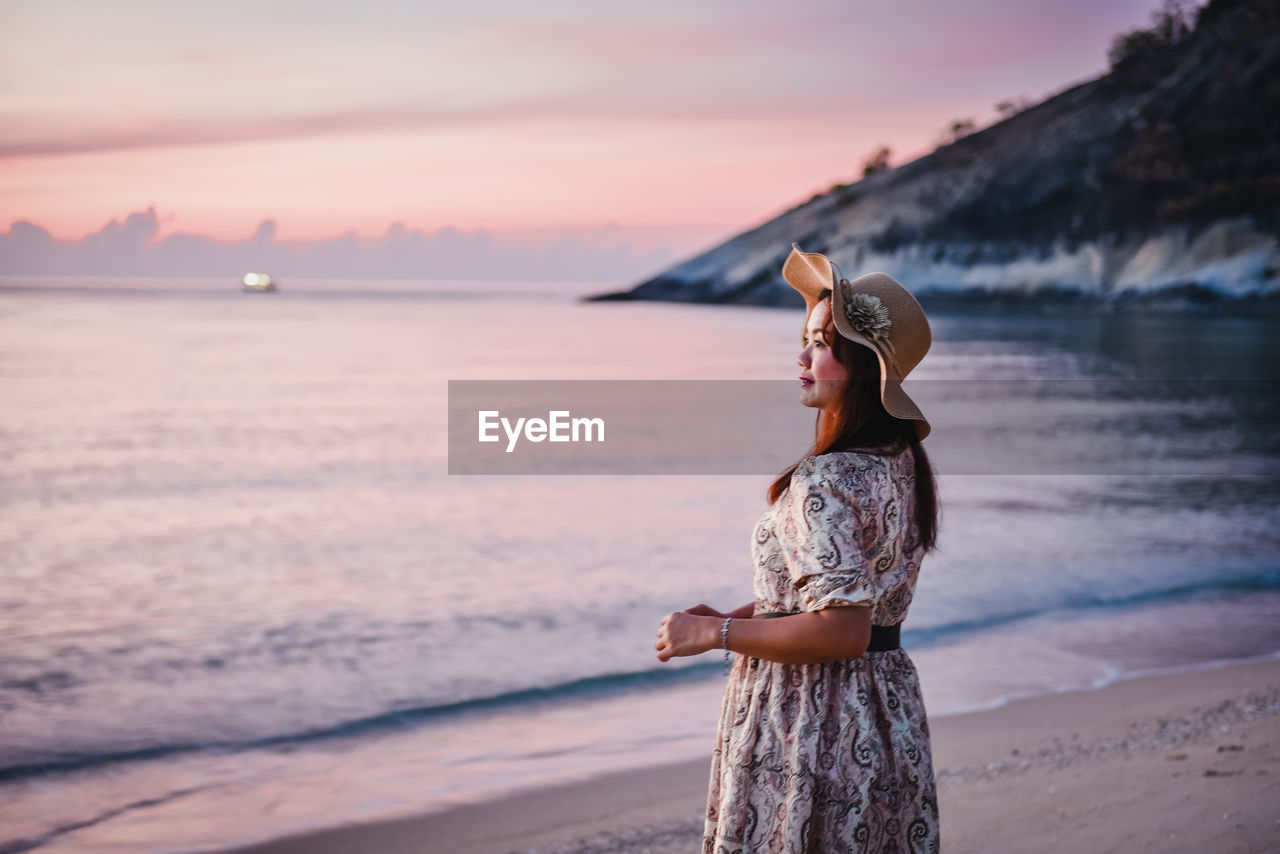 WOMAN STANDING ON BEACH AGAINST SEA DURING SUNSET