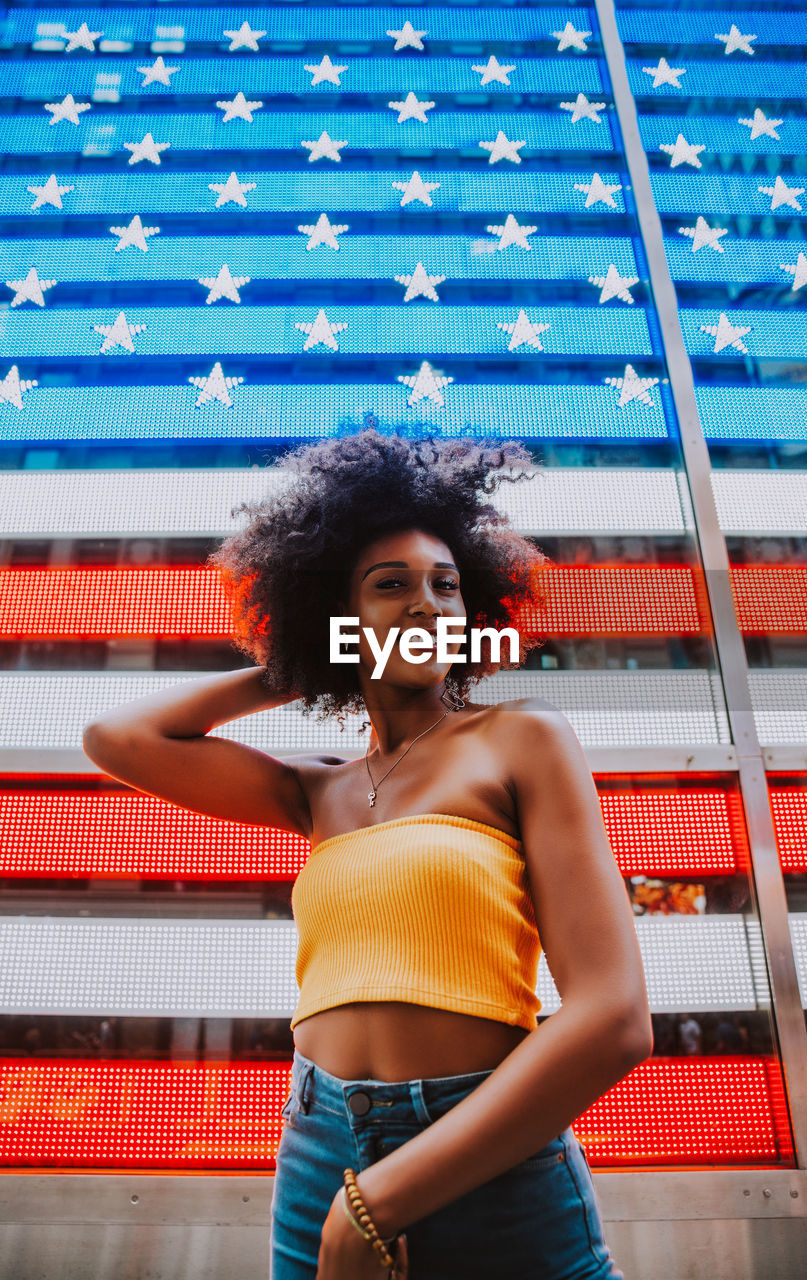 Low angle portrait of happy young woman standing against neon american flag in city