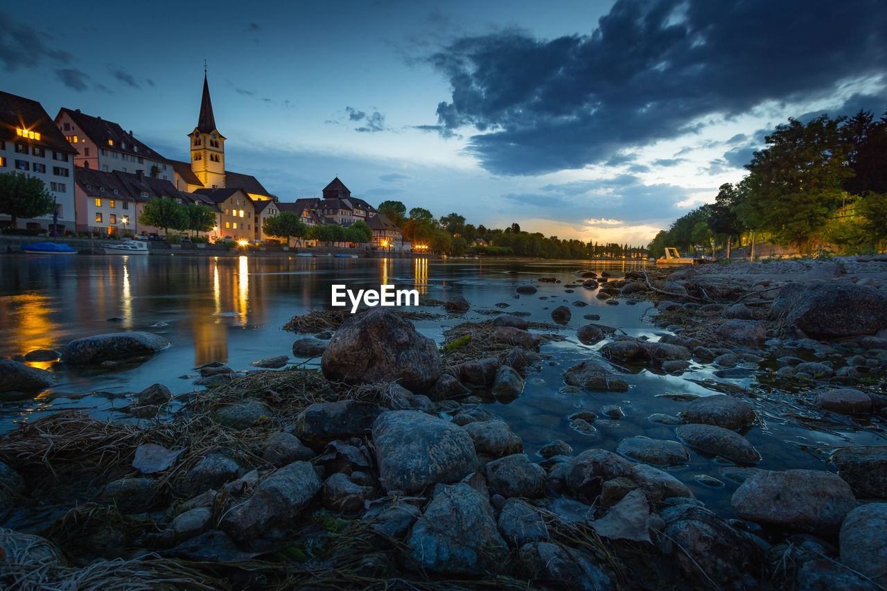 RIVER BY ILLUMINATED BUILDINGS AGAINST SKY AT SUNSET