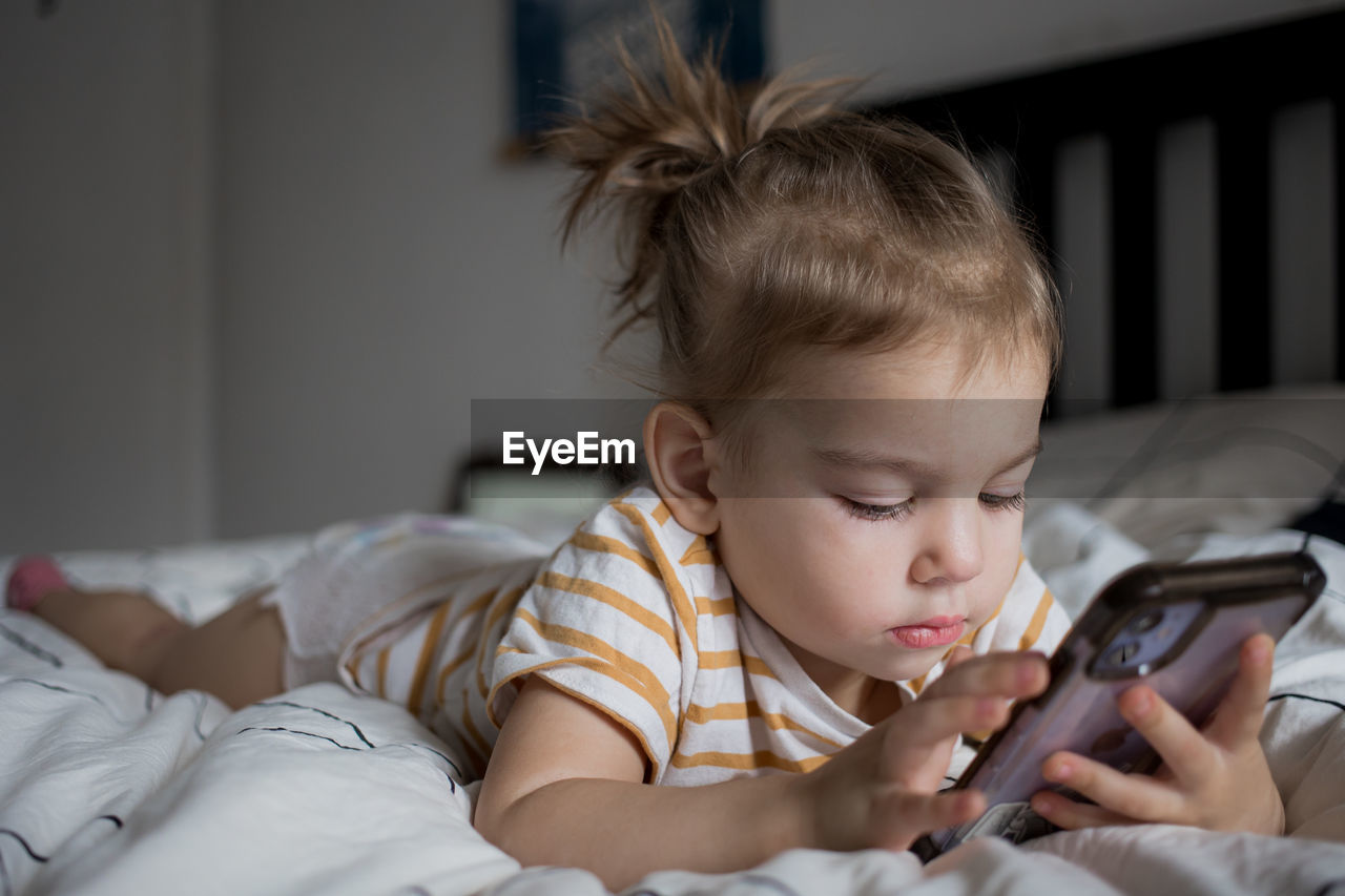 Little toddle girl playing with smartphone laying on the bed at home. caucasian toddler girl.