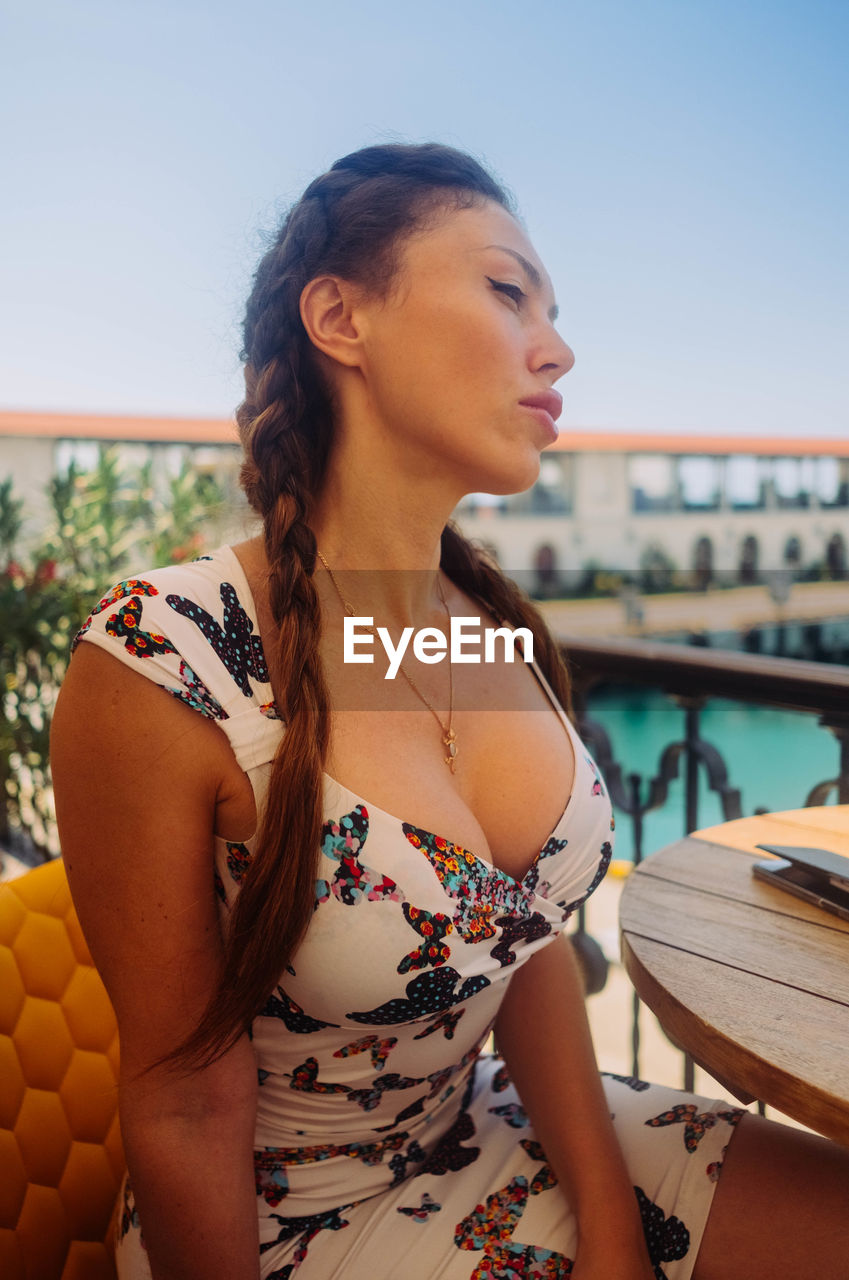 Beautiful woman looking away while sitting by railing against sky