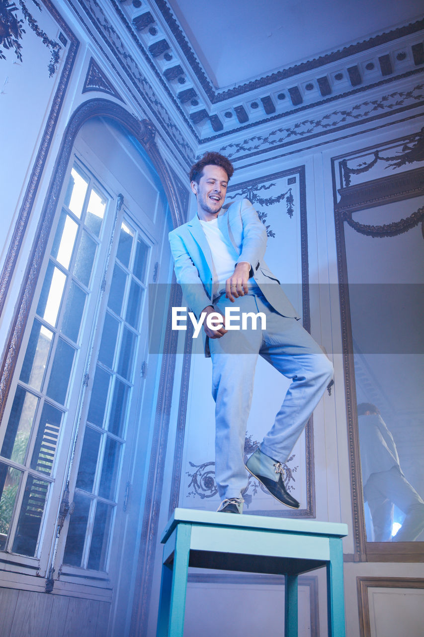 From below of expressive happy male in stylish suit balancing on table in posh room with blue neon light looking away