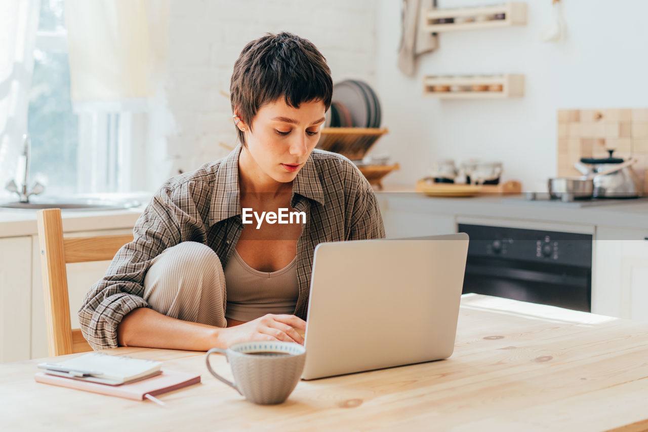 Young short-haired modern woman working on the computer while sitting at the table in the kitchen