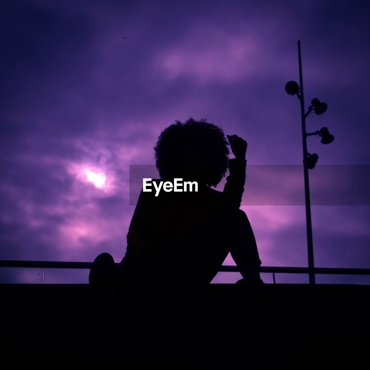 SILHOUETTE BOY SITTING ON RAILING AGAINST DRAMATIC SKY