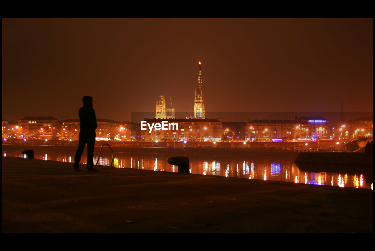 Silhouette person standing by river and illuminated industry at night