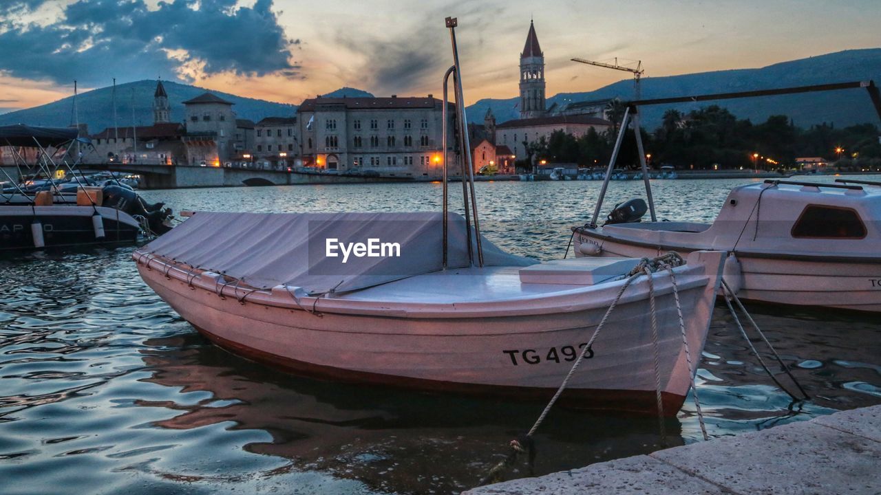 Sailboats moored on sea by city against sky during sunset