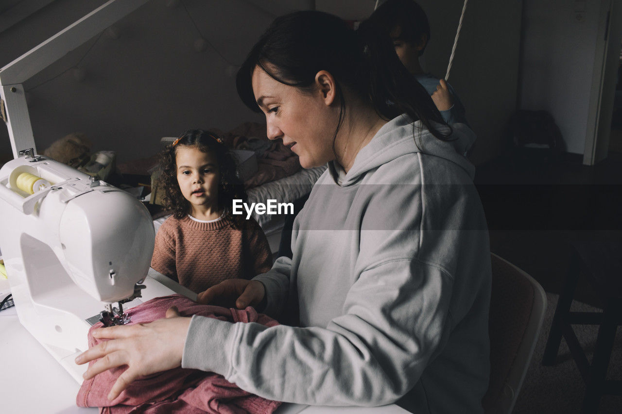 Daughter looking at mother sewing clothes at home