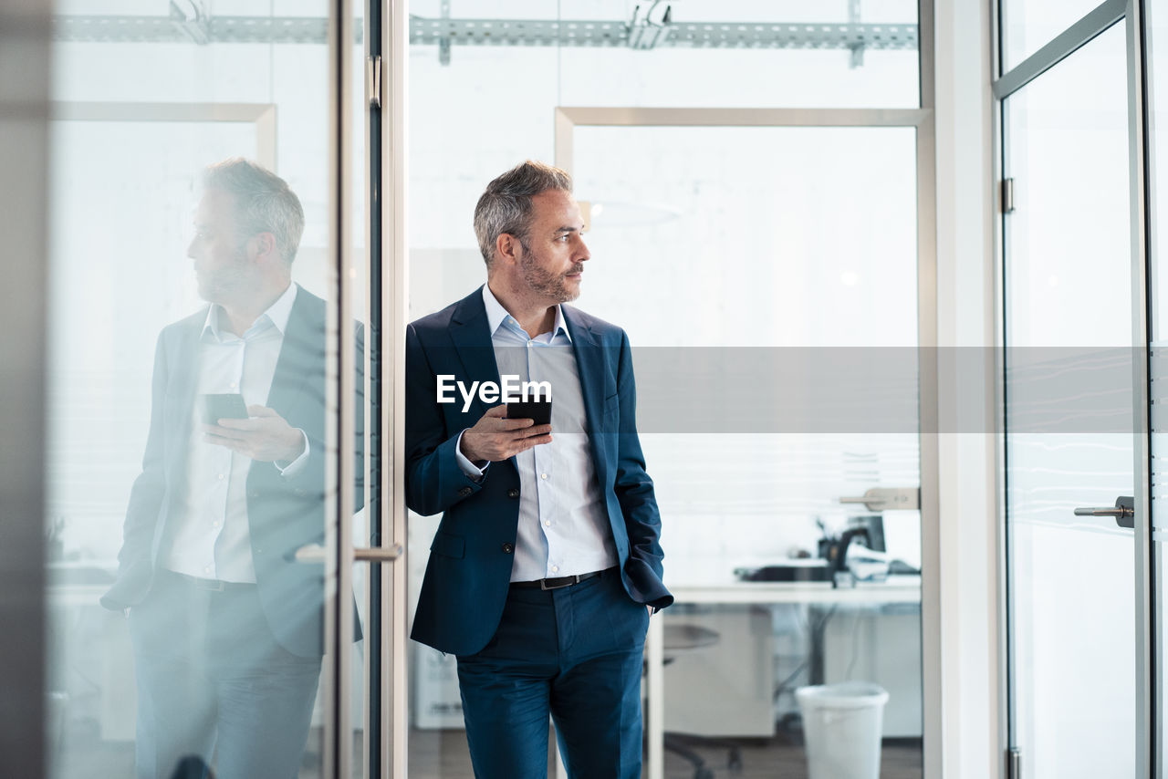 Thoughtful businessman leaning with hand in pocket holding smart phone at office