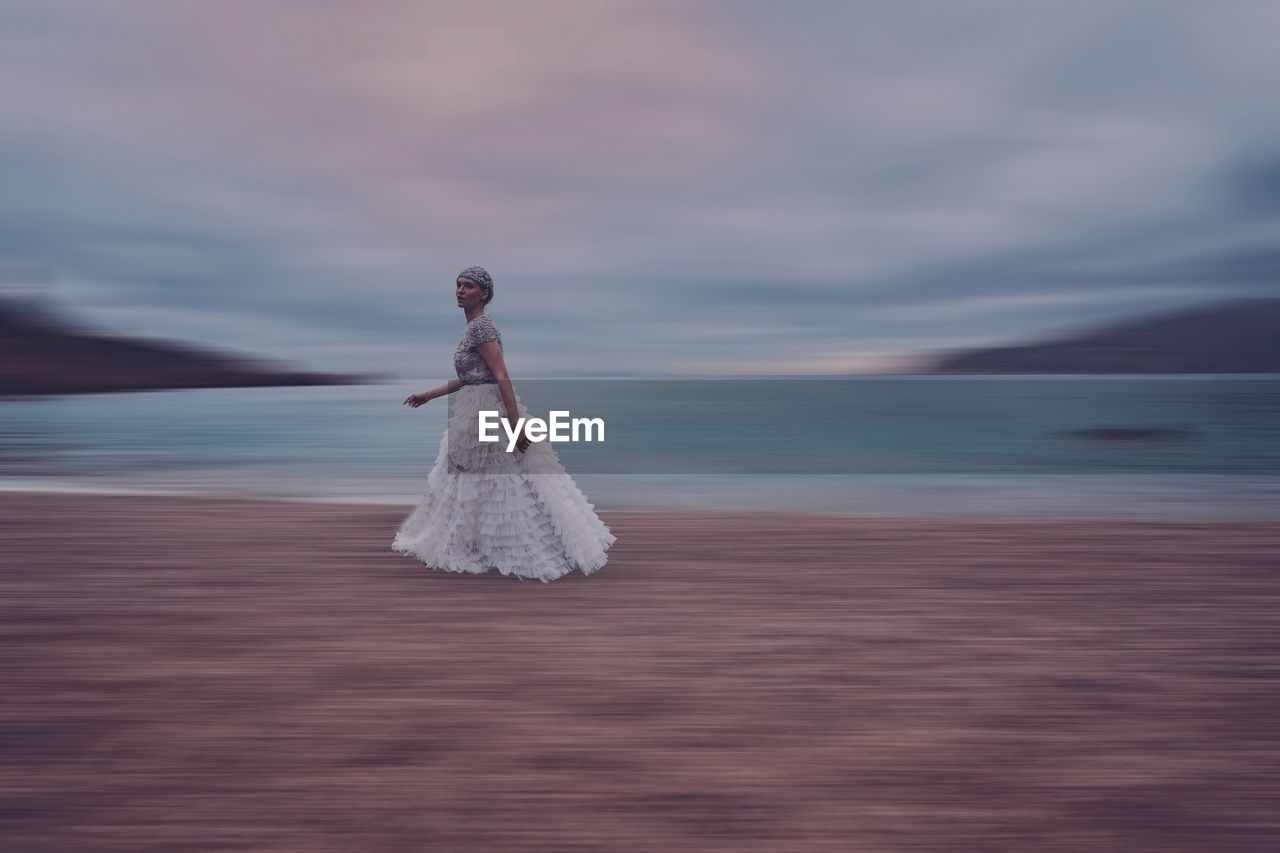 Blurred motion of woman walking at beach against sky during sunset