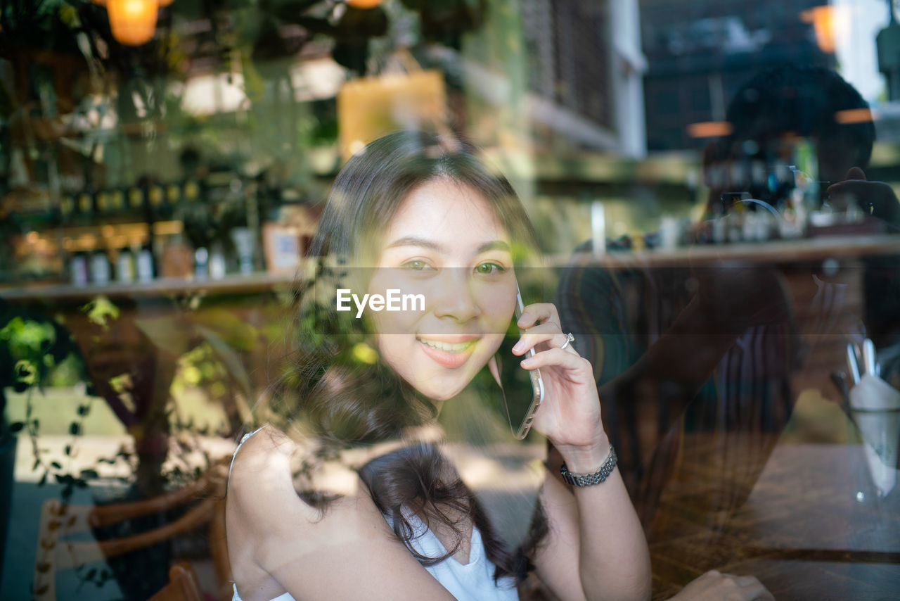 Portrait of woman using mobile phone while sitting at table seen through glass window