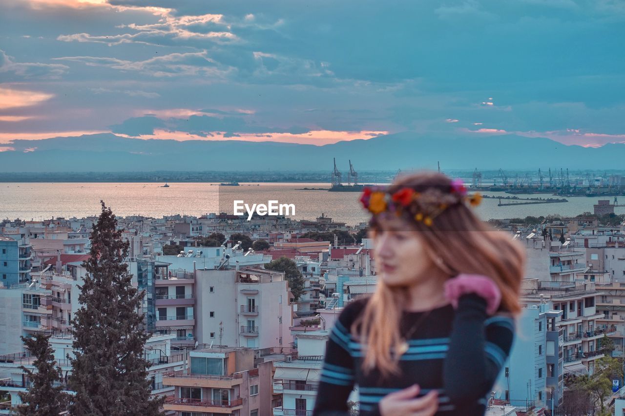 Woman standing by buildings against sea in city during sunset