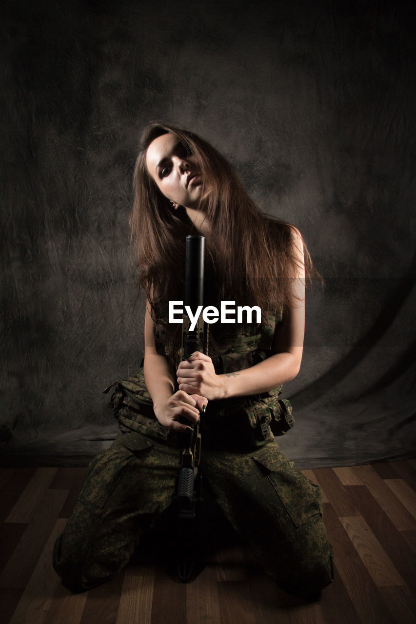 Portrait of young woman holding gun while kneeling against wall