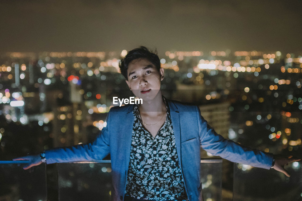 Portrait of young man standing against illuminated cityscape at night