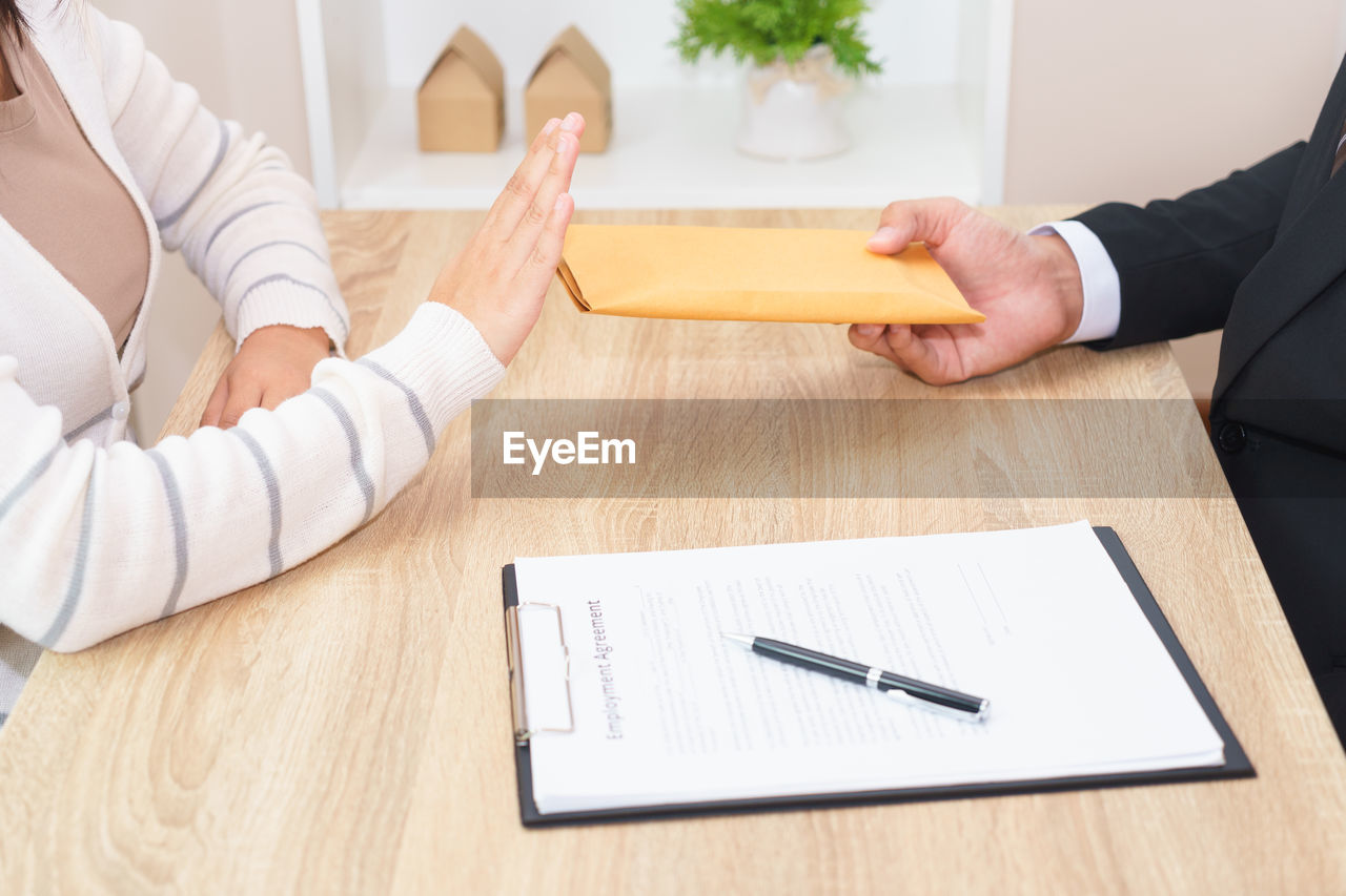 Midsection of woman showing stop gesture businessman giving envelop on office desk