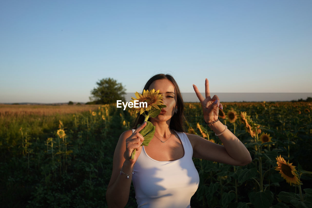 Portrait of young woman holding sunflower gesturing against sky during sunset