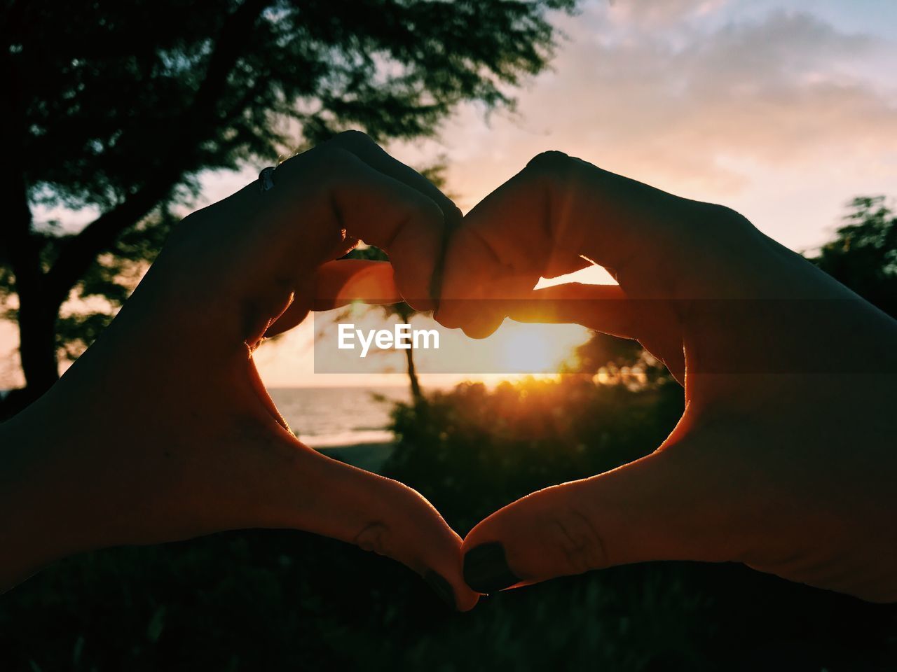 Cropped hands of woman making heart shape against sky during sunset