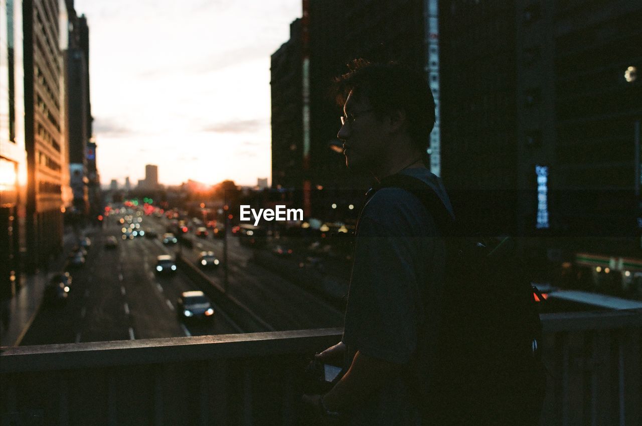 Man standing by railing on bridge in city during sunset