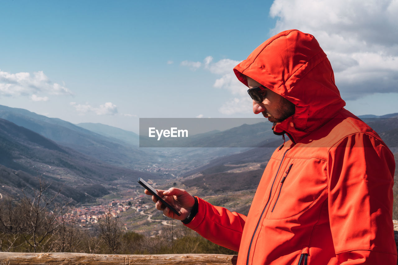 Caucasian male in a red coat typing with his smartphone with mountains in the background 