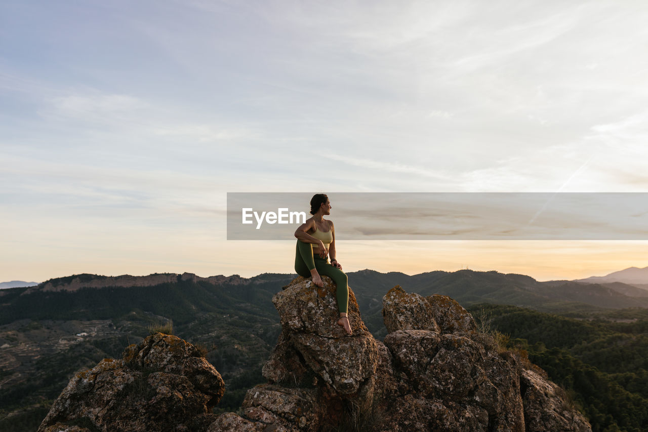 Young confident barefoot female in activewear relaxing on rocky cliff while having yoga practice at sunrise