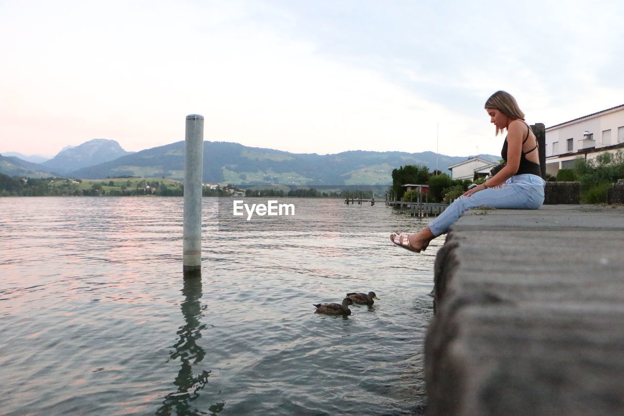 Full length of woman sitting on pier over ducks swimming on lake against sky