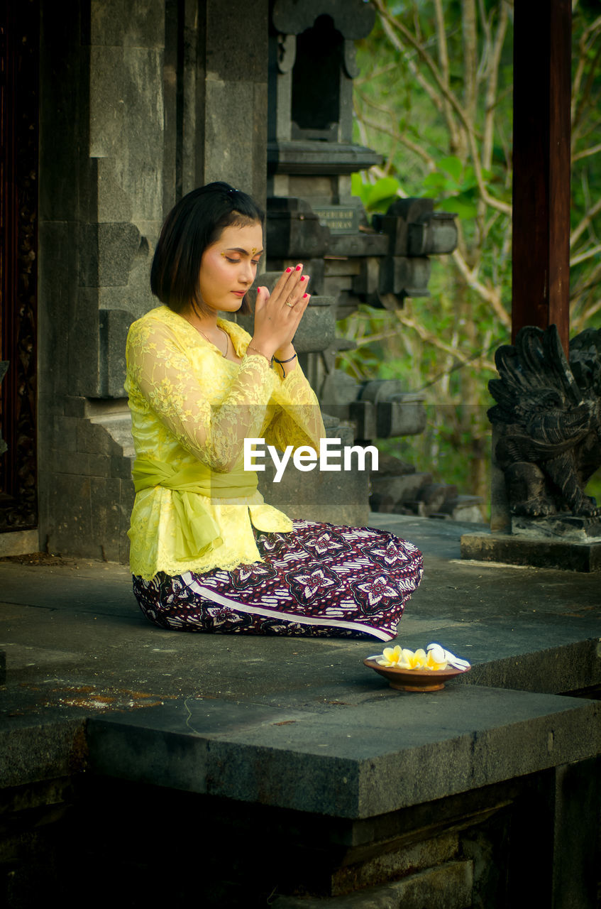 A woman wearing traditional clothing from bali is praying in front of hinduism temple