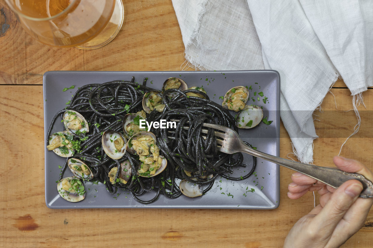 Female hand holds a fork with black spaghetti with clams. glass of white wine and linen napkin.