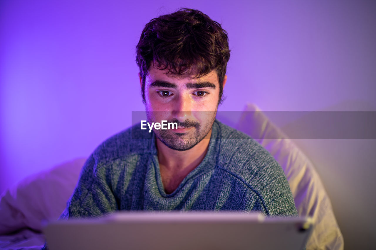 Man using digital tablet sitting on bed against wall at home