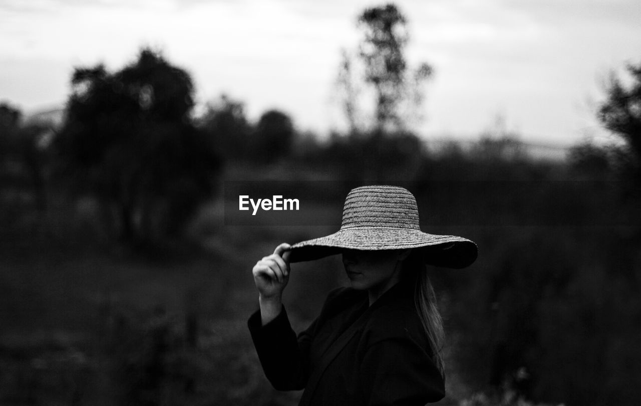 Woman holding hat while standing on field against sky