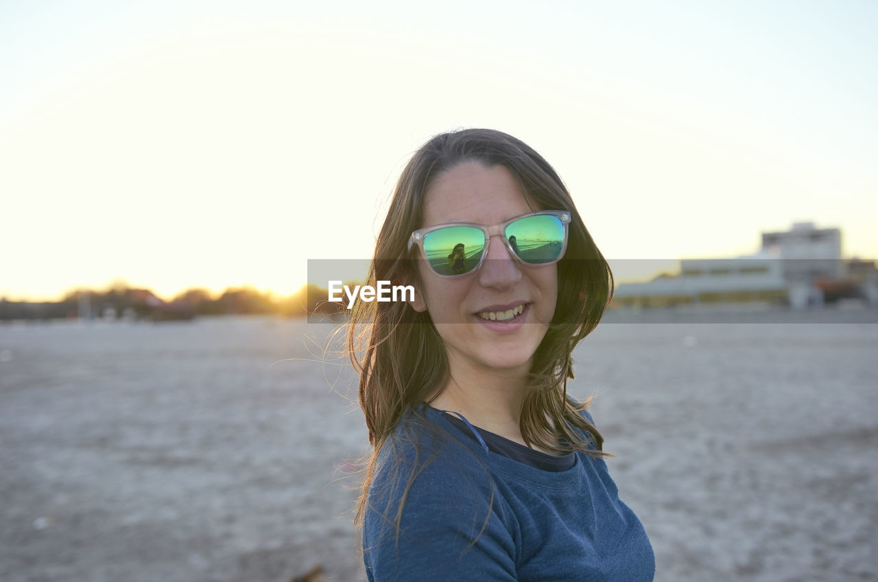 Portrait of woman wearing sunglasses while standing at beach against clear sky during sunset