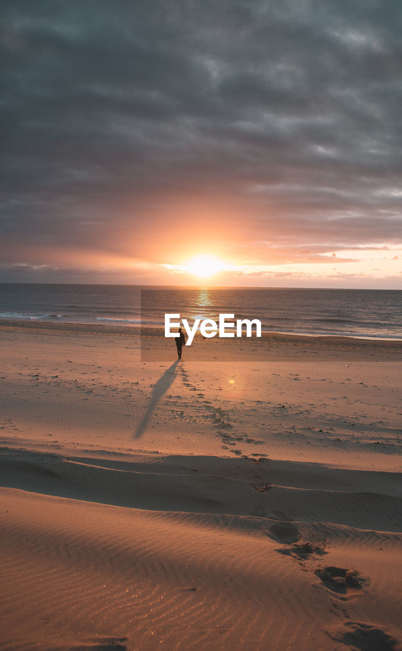 Silhouette man walking towards shore against cloudy sky during sunset