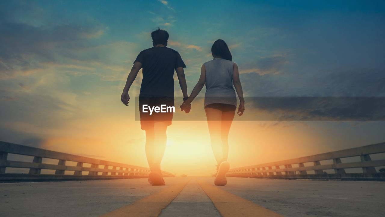 Rear view of couple holding hands while walking on bridge against sky during sunset