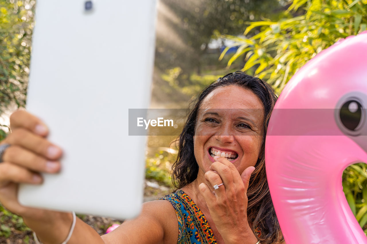 Beautiful smiling middle aged woman taking a selfie sitting on a pink flamingo shaped inflatable toy