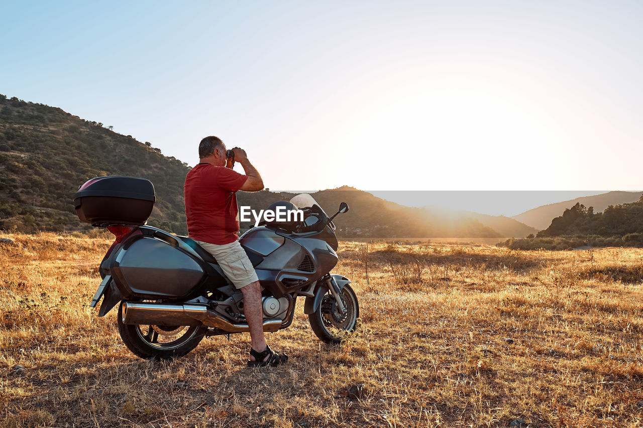 On the road.tourist man traveling on motorcycle, looking through binoculars at mountains in sunset