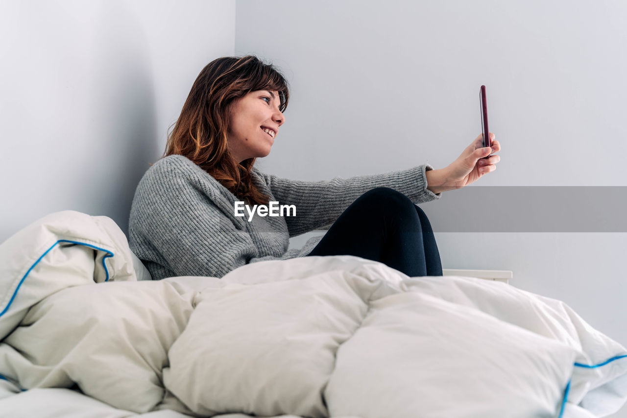 Side view of woman taking selfie with smart phone while sitting on bed
