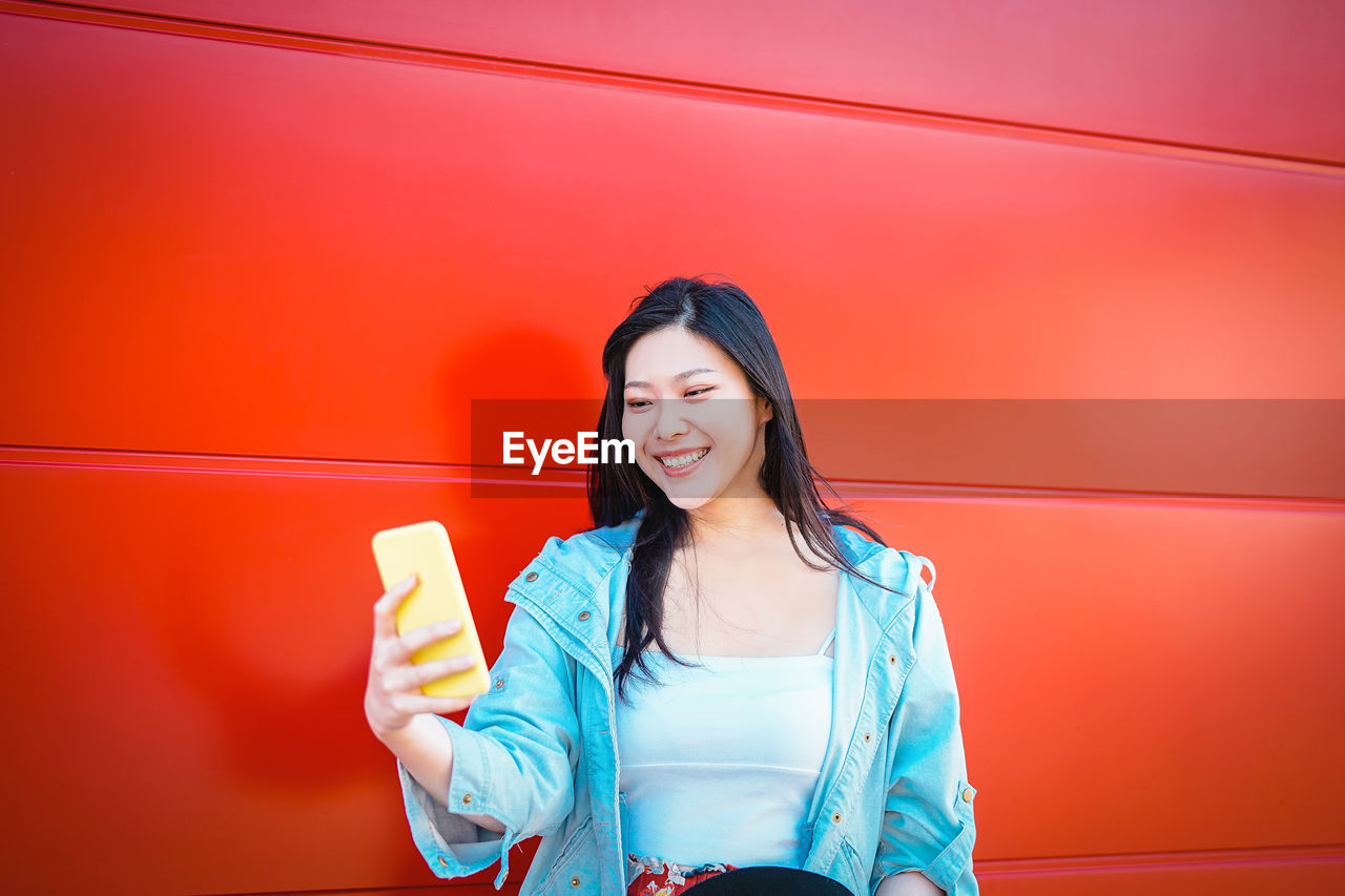 Smiling young woman taking selfie from mobile phone by red wall