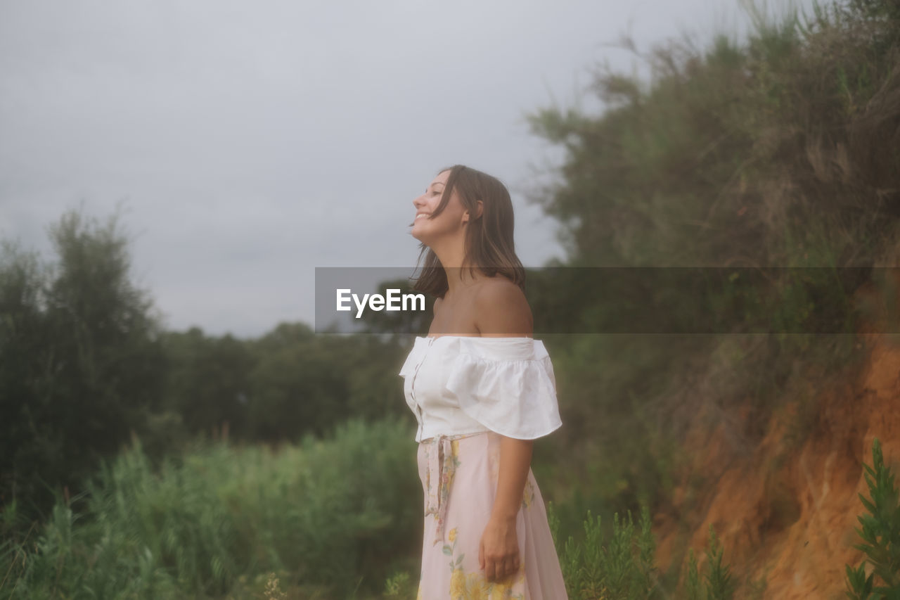 Side view of young woman standing amidst plants against sky in forest