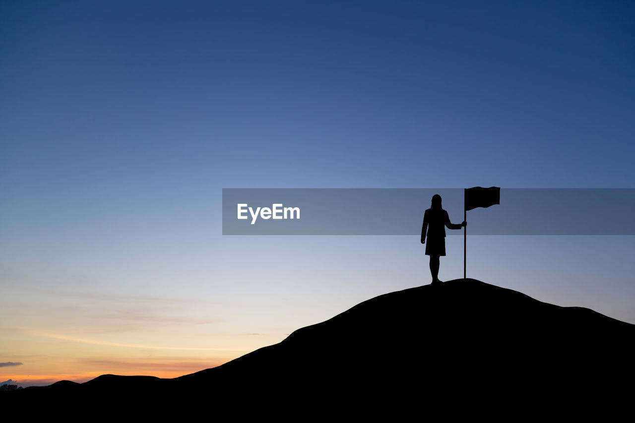 Silhouette person standing on mountain by flag against sky during sunset