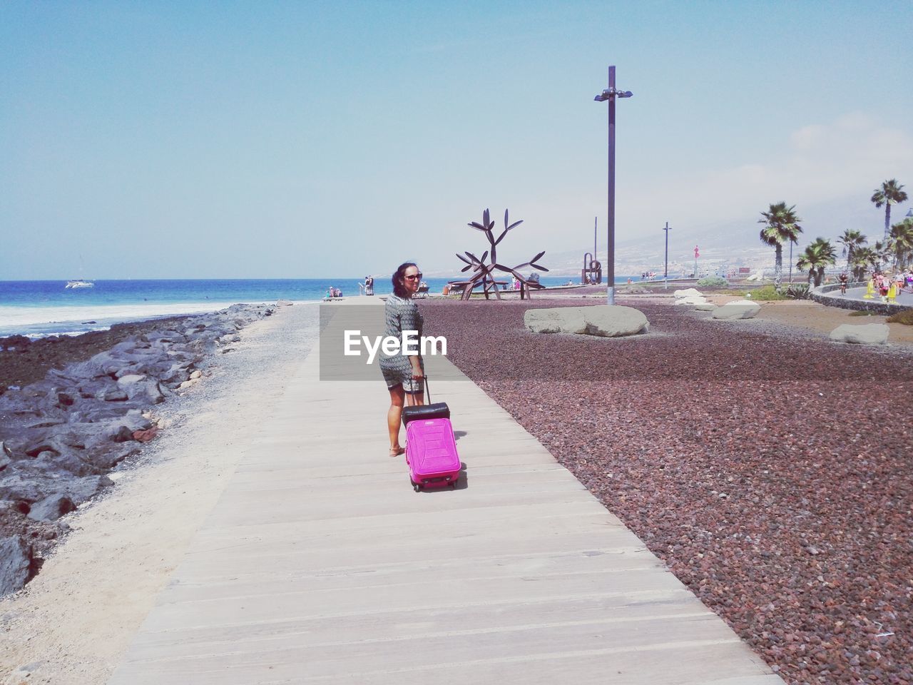 Full length of woman pulling luggage on footpath against sea during sunny day