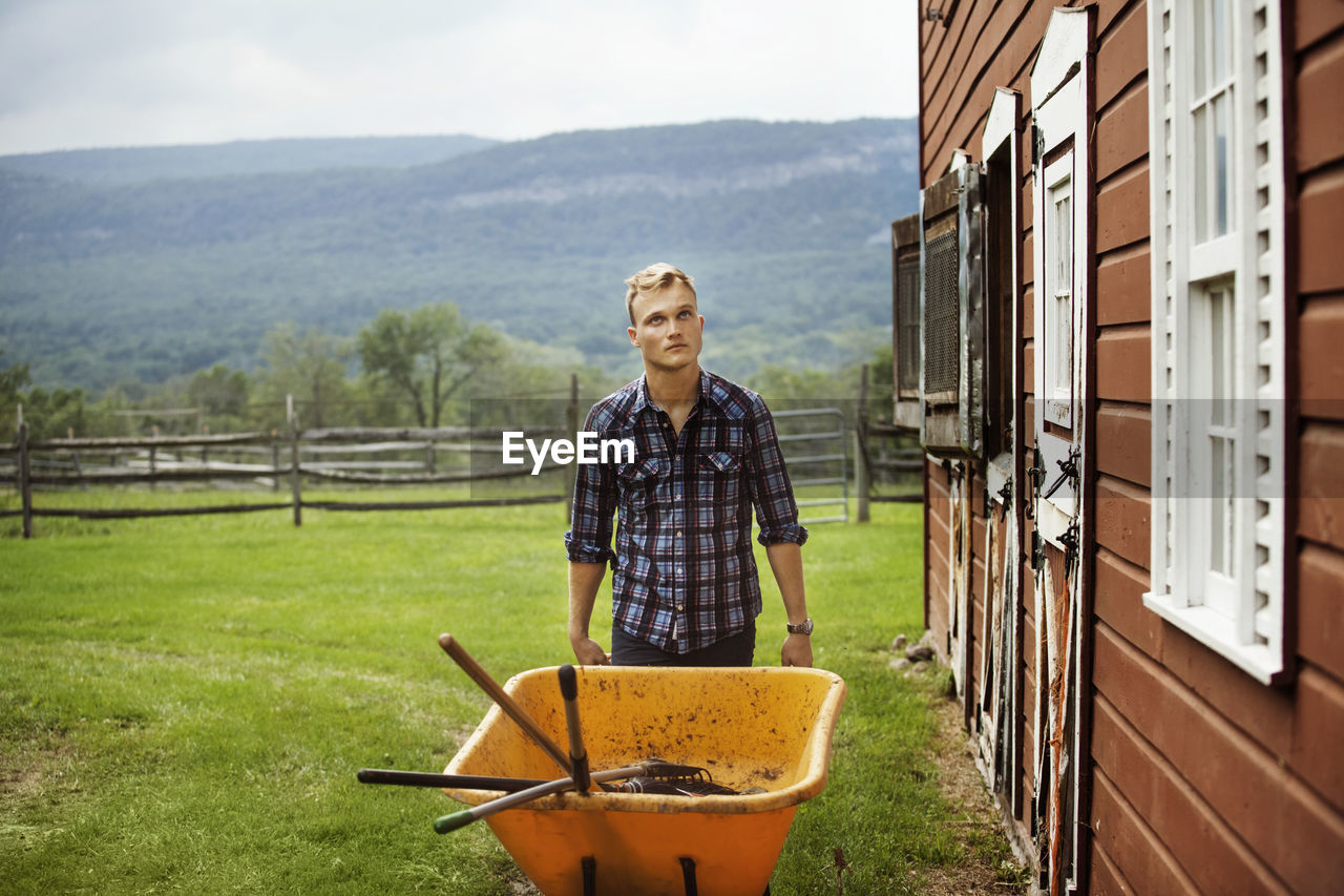 Rancher looking up while pushing wheelbarrow in farm against mountain