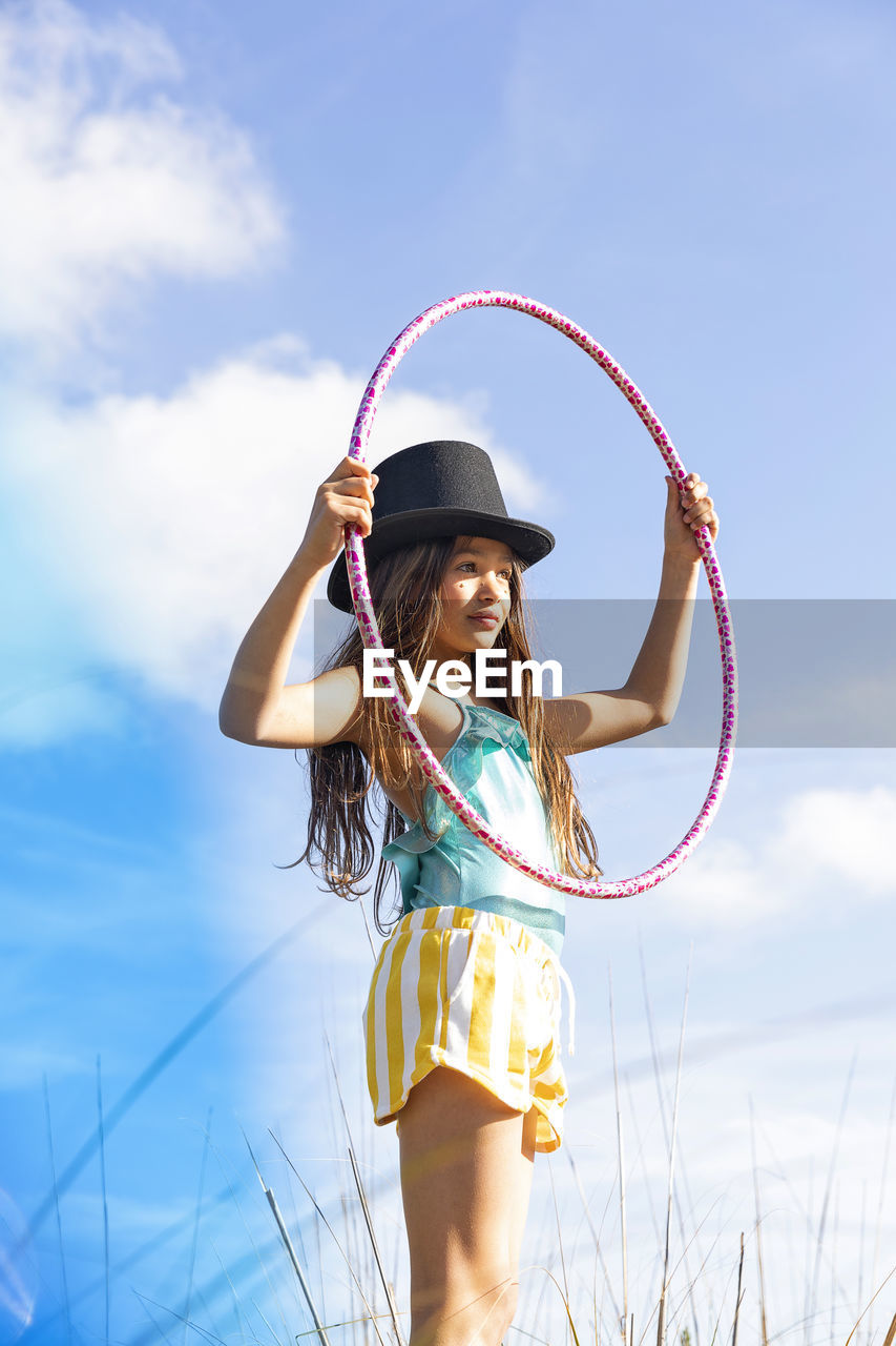 Girl wearing hat holding plastic hoop under sky