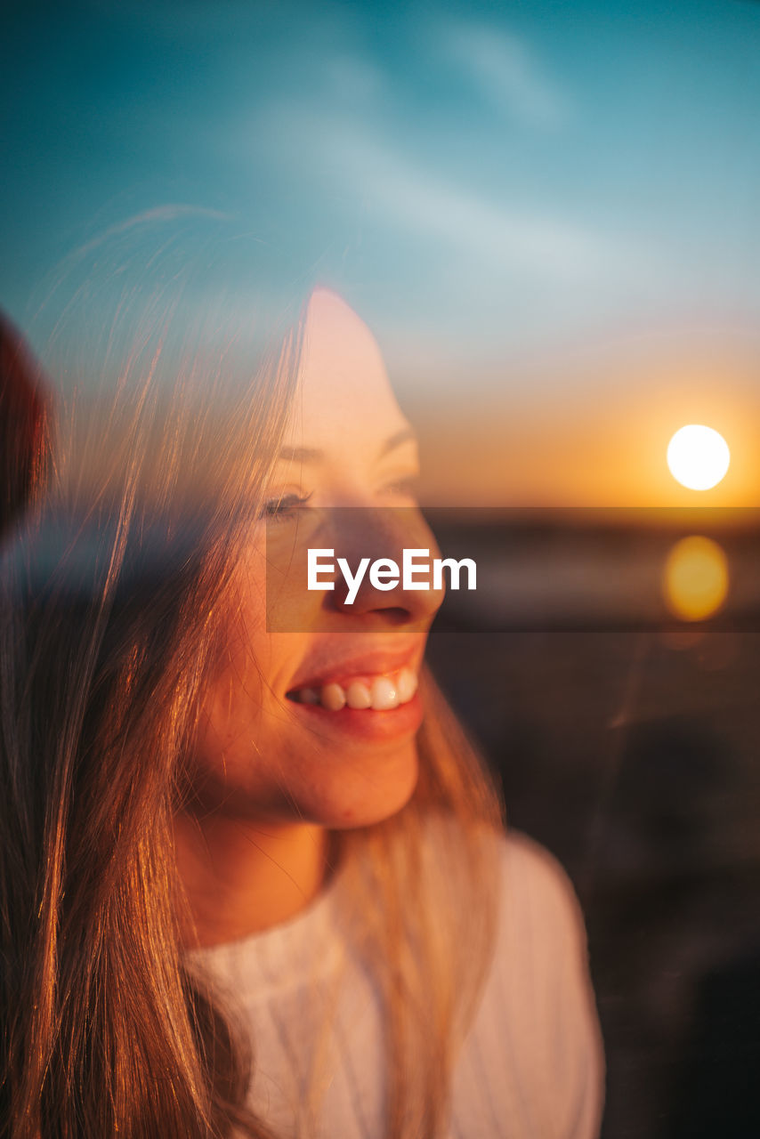 PORTRAIT OF A SMILING YOUNG WOMAN AGAINST SKY DURING SUNSET