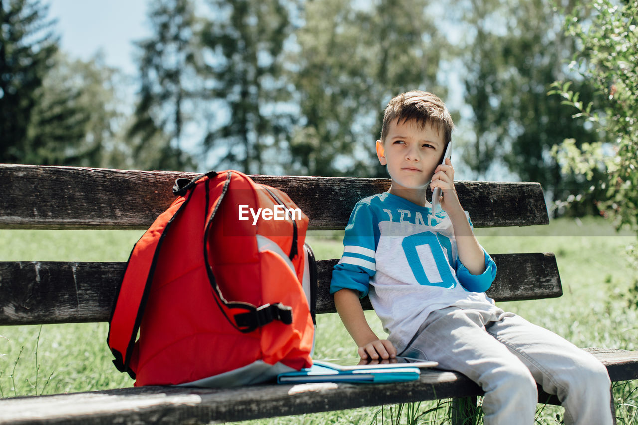 Boy using mobile phone while sitting on bench in park during sunny day