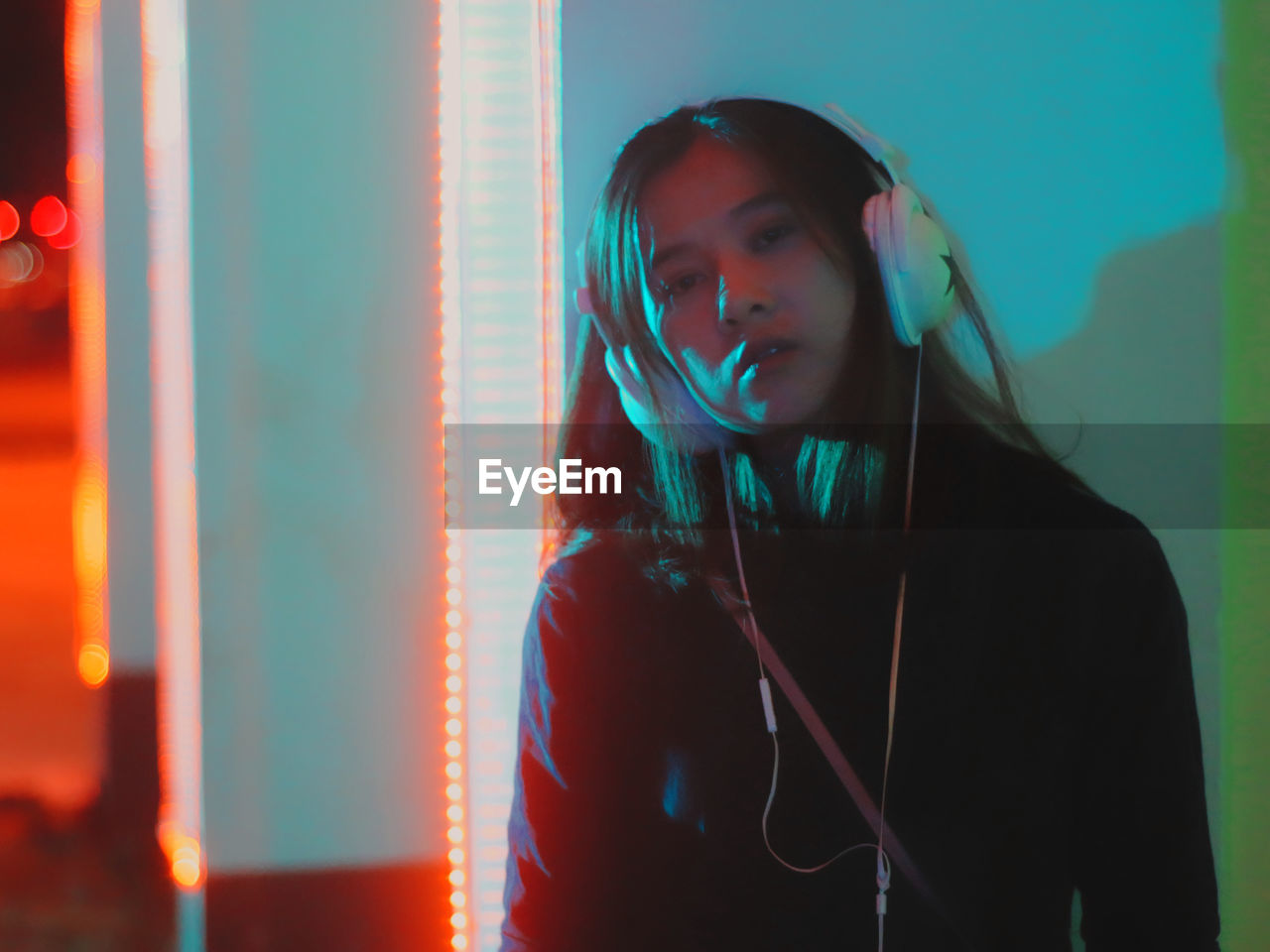 Portrait of young woman listening music against illuminated wall