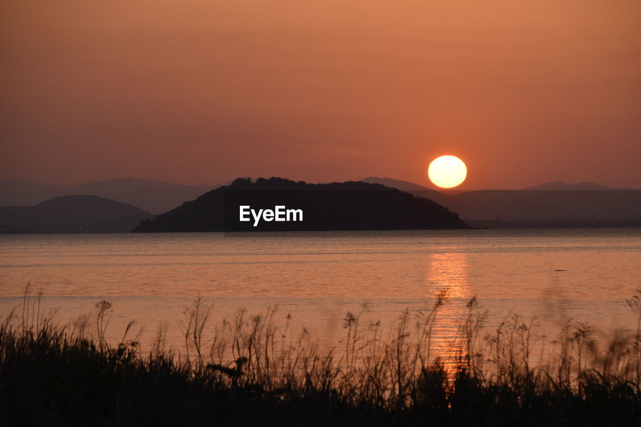 SCENIC VIEW OF LAKE BY SILHOUETTE MOUNTAINS AGAINST ORANGE SKY