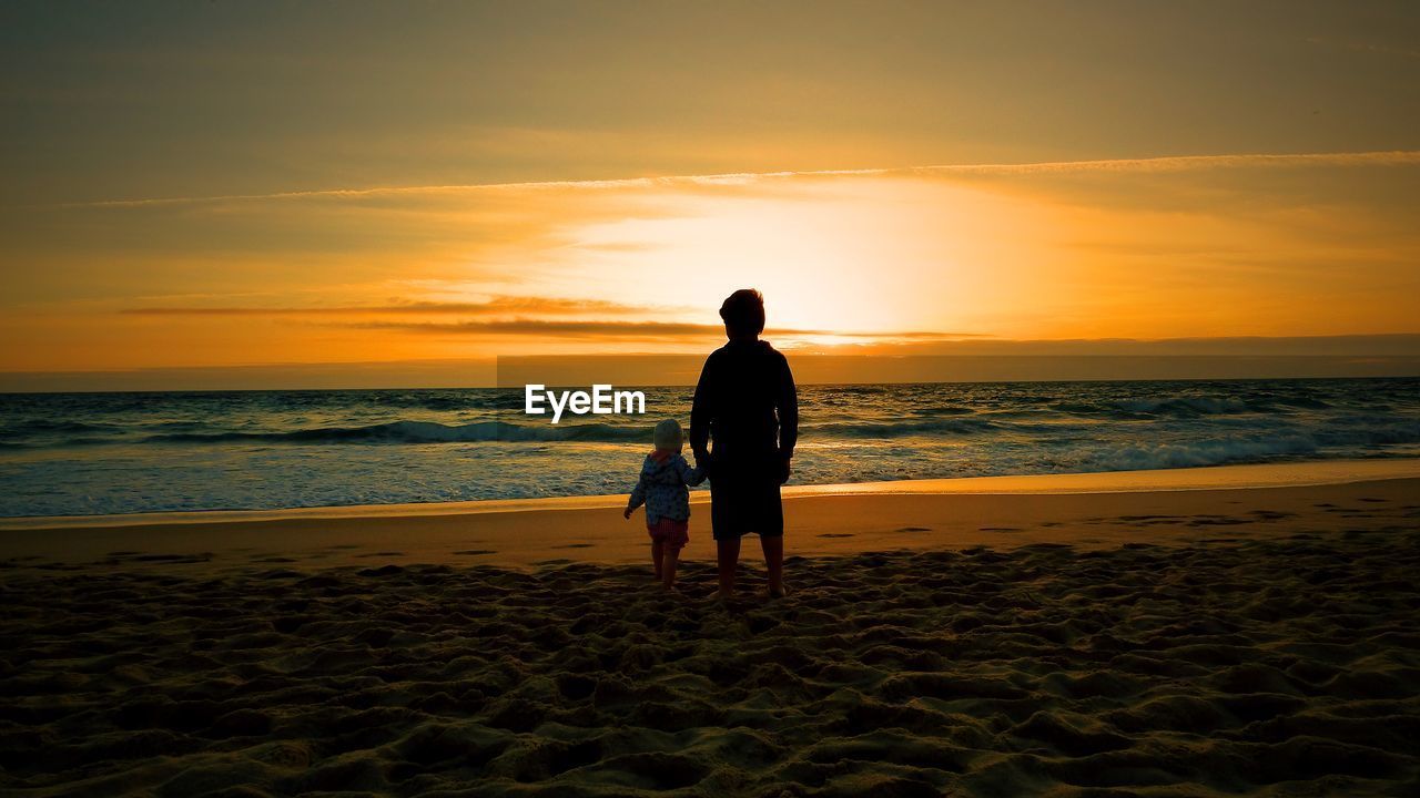 Silhouette man with son standing on sandy beach during sunset