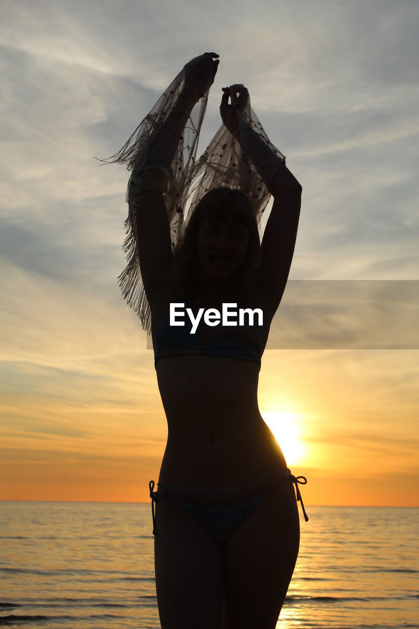 WOMAN WITH ARMS RAISED AT BEACH DURING SUNSET